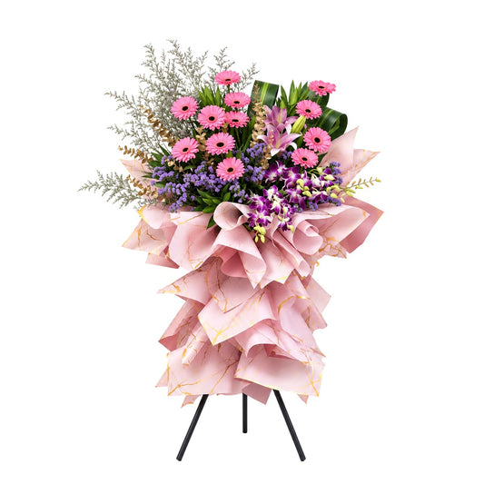 Blooming Success congratulatory flower stand with pink gerberas, pink lily, purple statice and orchids on a pink-gold wrap