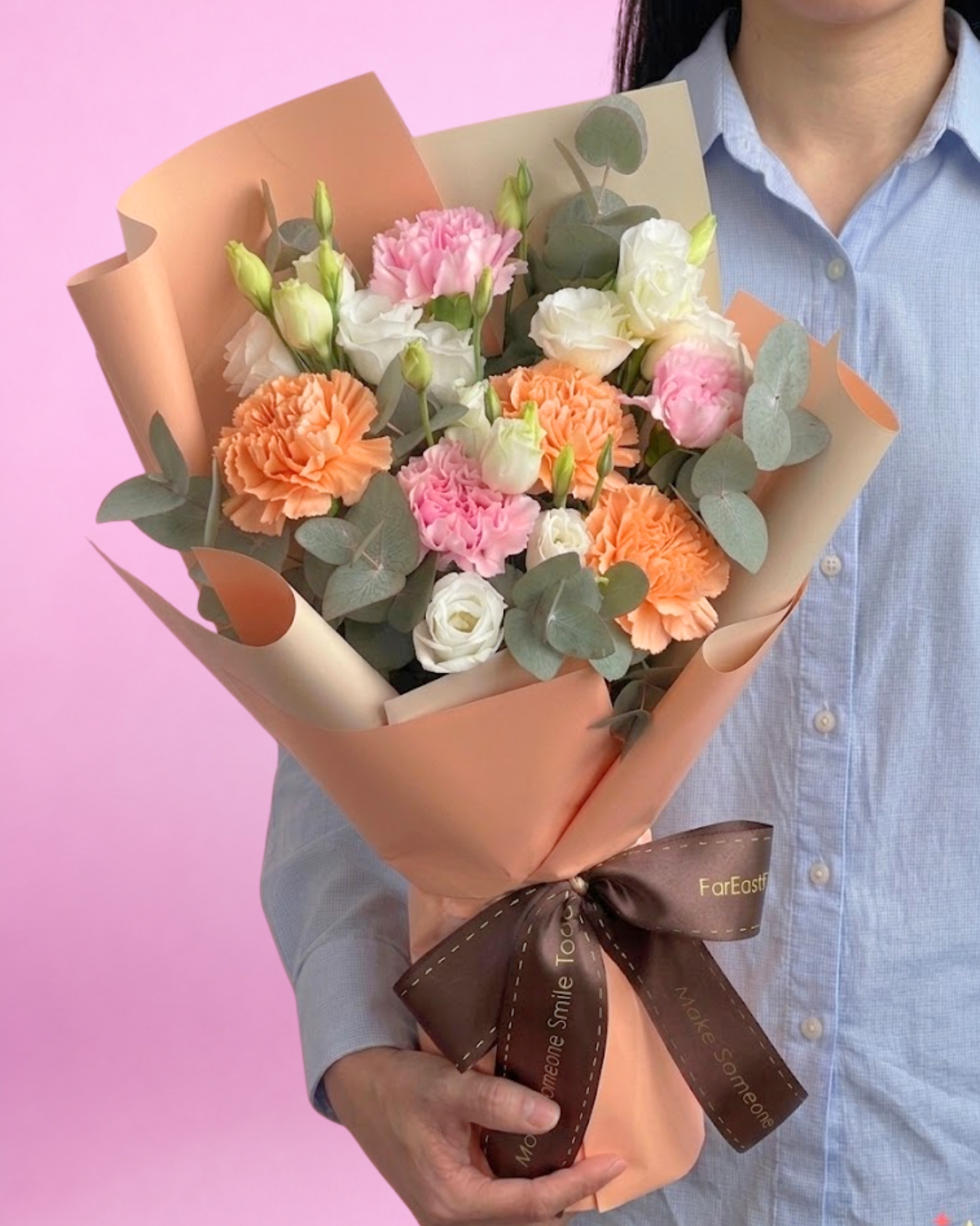 Bouquet of flowers wrapped in brown paper with a brown ribbon held by a person against a pink background