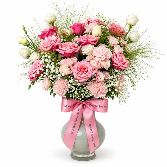 Blush Romance table flower arrangement with pink and cream roses, pink carnations, white eustoma and baby’s breath in a frosted glass vase with pink ribbon